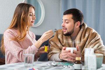 Fototapeta premium Young bearded man with symptoms of viral disease sitting at home table wrapped in plaid while worried wife trying to help him ..
