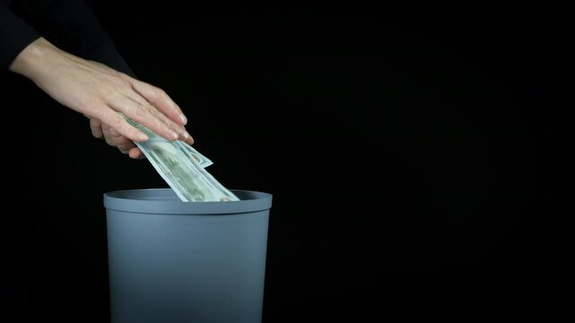 Dollars falls into trash at inflation. A view of woman hands put her money into trash bin on the black. A concept of world crisis problems.