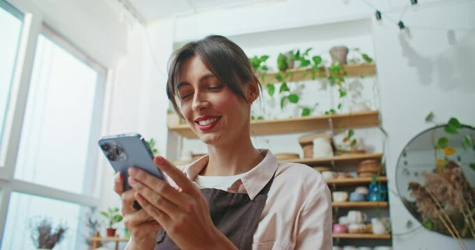 View from below of charming Caucasian woman actively typing on smartphone screen. Smiling after receiving funny message or positive reply. Getting distracted from work. Wearing apron.