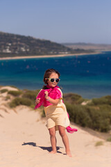 Beautiful girl in a yellow dress and a pink scarf walking on sand dunes, sea visible in the distance. Sunny summer day, beautiful sand dunes, wild sea beach in the background with many surfers