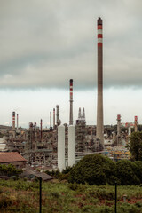 A vertical telephoto shot of an industrial oil refinery with multiple chimneys and cooling towers emitting fumes. Complex network of pipes, machinery, and structures, with greenery in the foreground