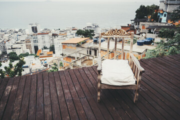 Wooden rocking chair with white cushion on a deck overlooking a hillside favela and urban buildings with an ocean view in the background. Rustic and serene setting