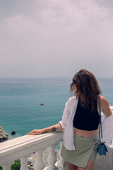 Fototapeta premium Beautiful girl posing on a balcony overlooking the sea. Girl in sunglasses and a white blouse enjoying a seaside vacation in Spain. Vacation at sea in the velvet season