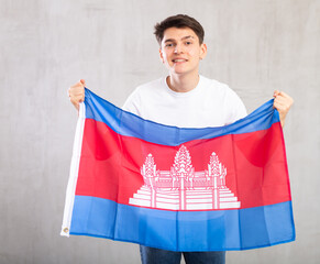 Enthusiastic young man holding large flag of Cambodia against light shadeless background