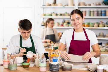 Fototapeta premium Joyous young man and woman in pinafores sculpting handmade jars of potter-earth material in art studio