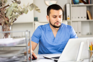 Obraz premium Skillful man physician sitting at table in his office and using laptop.