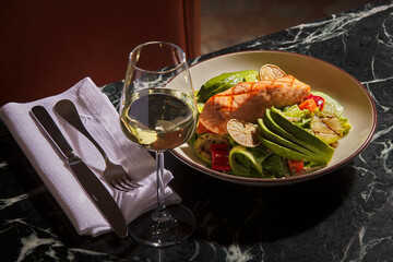 A plate of grilled salmon, avocado and salad with a glass of white wine. Luxurious and expensive gourmet meal on a black marble table.