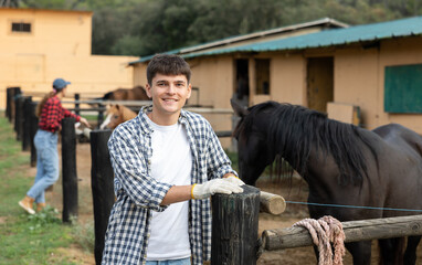 Male stable worker stands near wooden fence of paddock and watches horse. He is resting and taking...