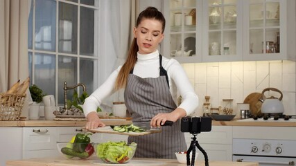 A woman in an apron slices vegetables in her bright kitchen, recording her cooking tutorial for viewers.