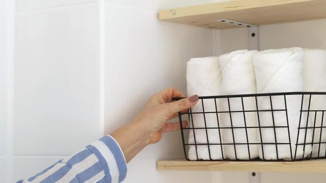 A woman folds white towels into a stylish basket on a shelf in the bathroom.