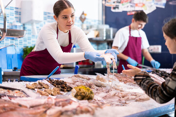 Portrait of friendly female fishmonger showing fresh fish to woman behind counter of seafood store