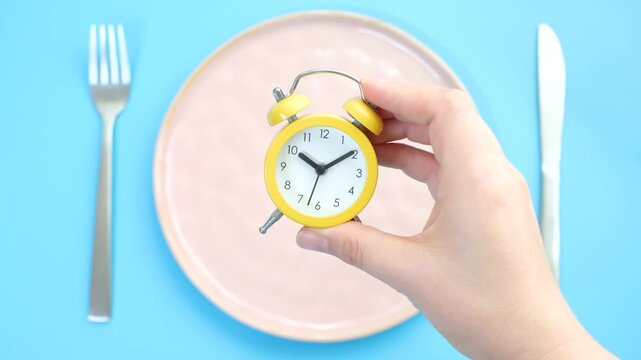 Female hand puts an alarm clock on an empty plate on blue background, top view.