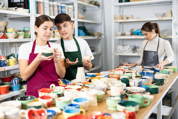 Smiling young man and women in pinafores examining handmade jars of potter-earth material in art studio