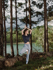Young woman practicing yoga in forest, balancing on one leg with hands raised, serene expression Nature and wellness concept