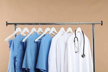 Different medical workers' uniforms and stethoscope on clothing rack against beige background