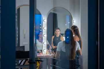 Seen through a window, makeup artist and model are reflected in mirrors illuminated by bright...