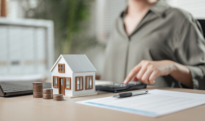 residential, rental, finance, wealth, calculator, agreement, mortgage, property, real, rent. A woman is sitting with calculator and house model. She is pointing at the calculator and the house model.