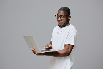 African American man surprised while using a laptop with glasses in a casual white t shirt Background is a plain gray color, conveying a modern and minimalist concept of technology