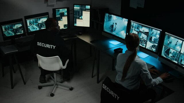 Full high-angle shot of two multiethnic security officers working in control room, Caucasian female guard noticing suspicious activity on CCTV cameras, discussing with African American male colleague