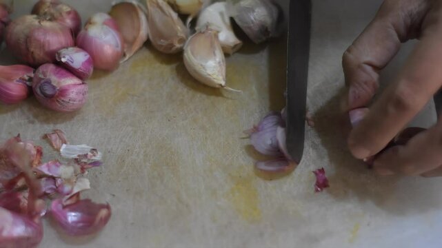 A close-up of a woman's hand cutting an onion on a white cutting mat, with a garlic and another onion placed beside. The scene focuses on precise food preparation in a clean kitchen setting.