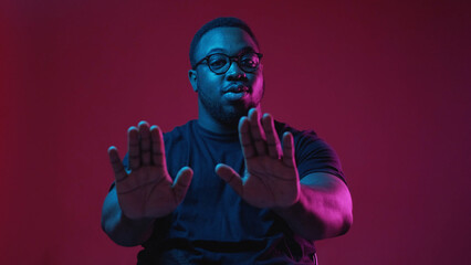 Stop gesture. Rejection refusal. Serious man with negative decline denial no hand sign in blue neon light isolated on dark burgundy background.