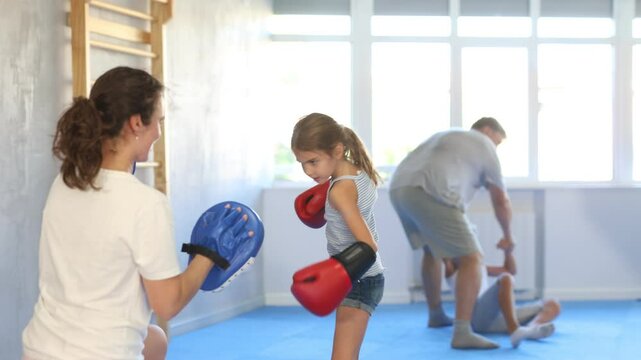 Girl and mother are boxing in gym, mom helps daughter child to work out force of blow. Modern positive family chooses active hobby