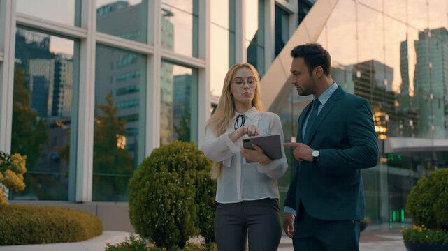 Two young successful Architects male and female business people outside big office building at sunset using Digital tablet. Collegues talking about Construction Project Outdoor. Teamwork
