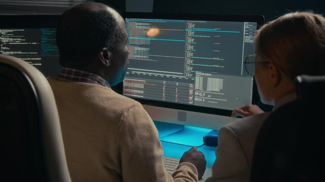 Medium rear shot of African American senior male programmer and Caucasian female junior colleague sitting together in front of screens, discussing structure of computer code
