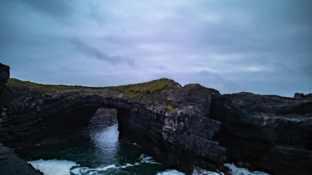 Timelapse of blue hour waves as ocean water crashes and rises below natural sea rch at Loop Head Kilkee, light fades over rugged coastal cliffs as clouds race in sky