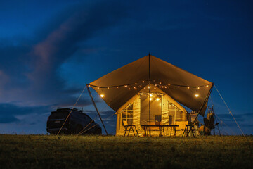 A tent is set up in a field with a car parked nearby. The tent is lit up with lights, creating a cozy and inviting atmosphere