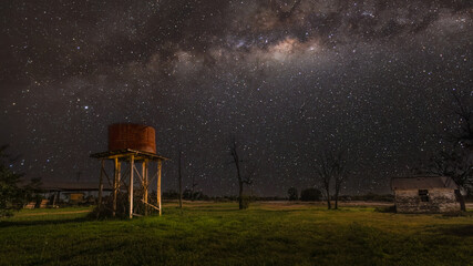 A lit up old corrugated iron water tank on timber poles and a deserted old building under the magnificent Milky Way Galaxy core in a night scene at Yelarbon in Queensland, Australia. © Shirley and Johan