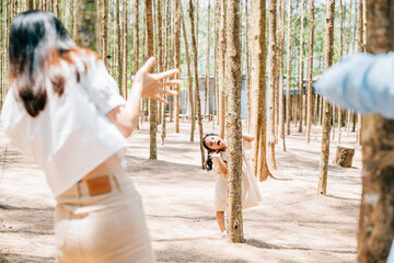 Asian family father and mother playing hide and seek back tree with daughter on forest nature park, happy parents funny together, Family enjoying moment of holiday