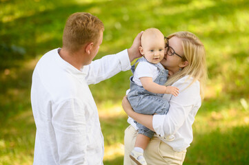 Fototapeta premium Mom tenderly holds baby in arms, showing affection, care, enjoying moments spent together outdoors and motherhood, dad tenderly strokes baby head. Happy family concept. International Day of Families