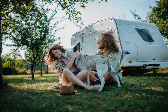 Young couple is on a camping trip in nature playing with dog, sitting in front of caravan. Caravan traveling for young people.
