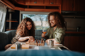 Fototapeta premium Young couple is on a camping trip in nature, sitting in caravan and playing chess. Caravan traveling for young people.