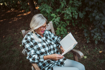 Beautiful older women is sitting in rocking chair in the garden and reading book. Female senior is enjoying a peaceful moment at home, savoring her freedom.