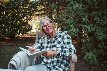 Beautiful older women is sitting in the garden and writing in her diary. Female senior is keeping...