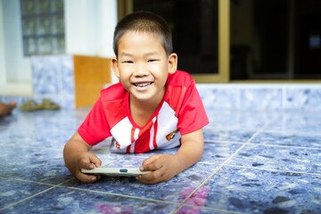 Close-up of a smiling red shirt boy looking at the camera while lying down and playing a game on his smartphone. © CW Photography
