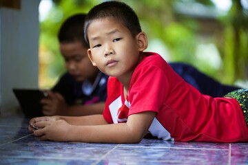 Close-up of a red shirt boy looking at the camera while lying down playing a game on his smartphone. - Powered by Adobe