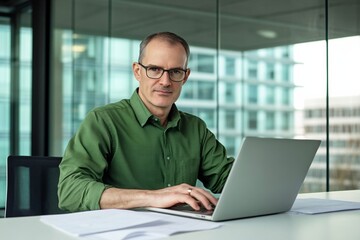 Fototapeta premium Middle-aged man with glasses working on a laptop at an office desk, blurred glass wall background, wide-angle professional photograph.