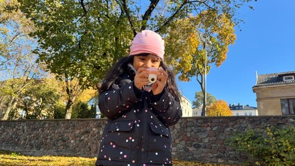 Beautiful Indonesian little girl holding mini kid camera in autumn season wearing padded jacket and pink beanie. Low angel