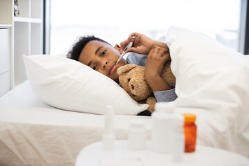 African boy sick with flu lies in bed measuring temperature with thermometer. Young child holds...