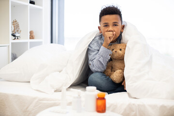 Sick African boy sits in bed and coughs holding teddy bear. Room is cozy with daylight streaming...