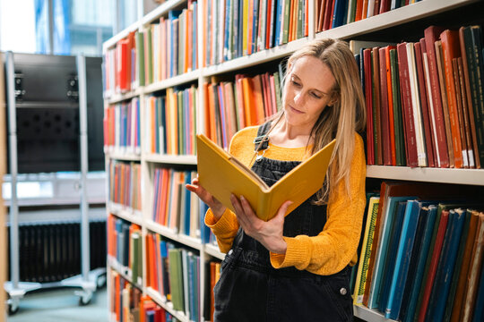 Librarian reading book and standing by bookshelves in library