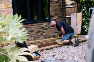 Mature workman kneeling on wooden planks near house