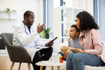 African male doctor speaks to concerned mother and son during examination. Mother and son, both of...