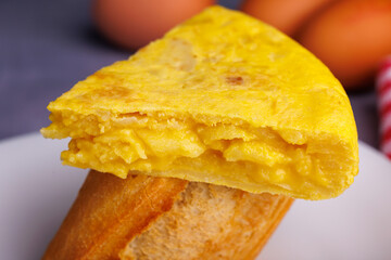 Close up of a Spanish omelette skewer with a bread portion, with eggs in the background, on a blue concrete table. Overhead view