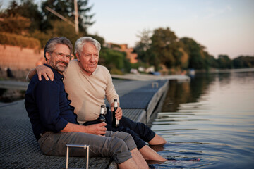 Smiling senior friends sitting with beer bottles on jetty