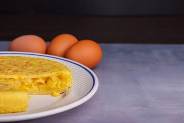 Traditional Spanish omelette with a missing piece, bread, eggs, and a red checkered napkin on a blueish concrete table