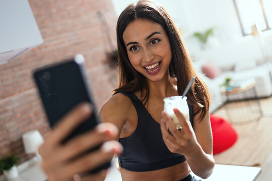 Young woman with yogurt jar taking selfie on mobile phone at home